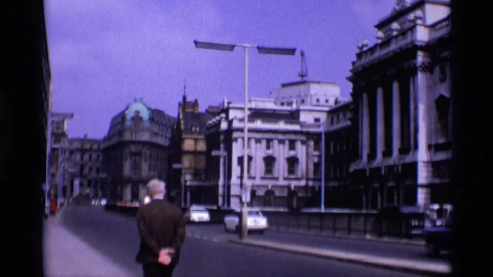 A 1969 London street view shows an adult man from behind, classic cars and prominent civic-looking buildings under a blue-purple sky.