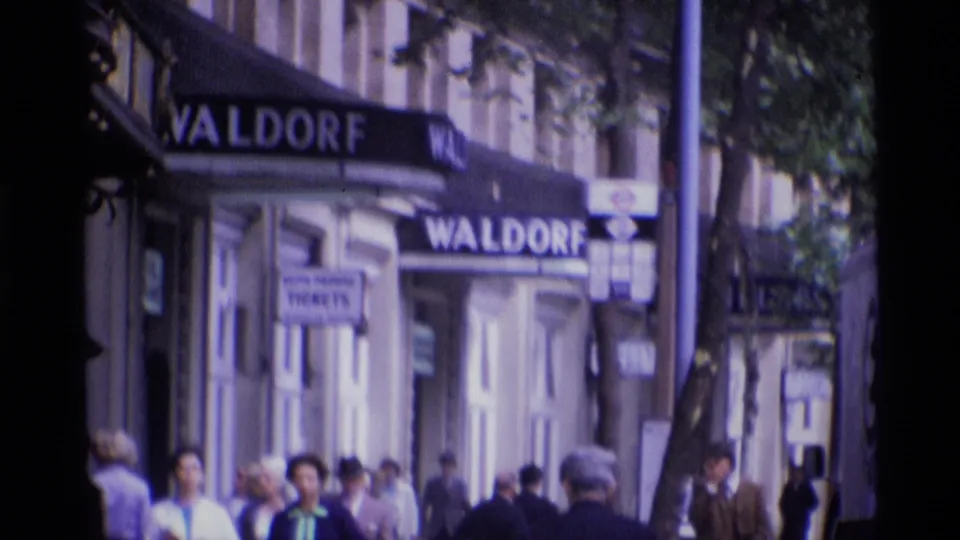 Pedestrians pass beneath Waldorf awnings on Aldwych in a soft, handheld 1969 London street view.