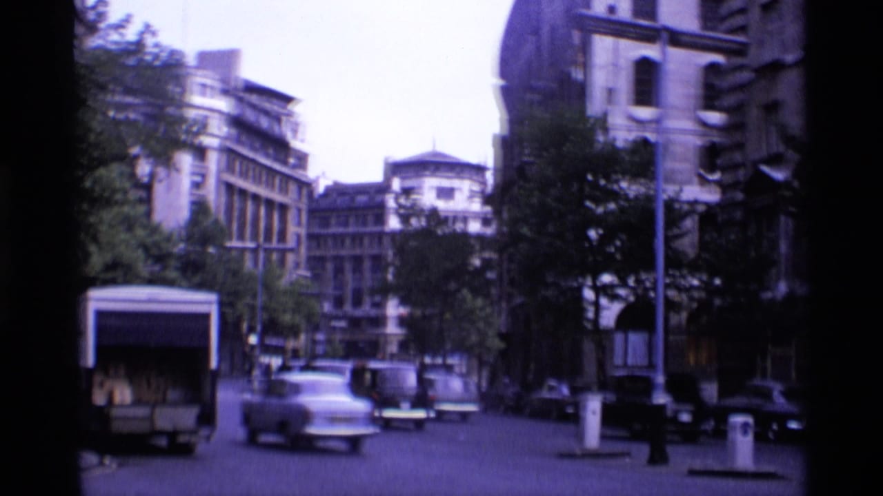 1969 London street view with classic cars moving past trees and older multi-story buildings in soft 8mm color.