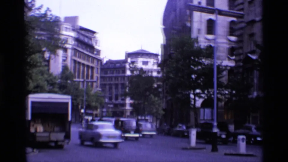 1969 London street view with classic cars moving past trees and older multi-story buildings in soft 8mm color.