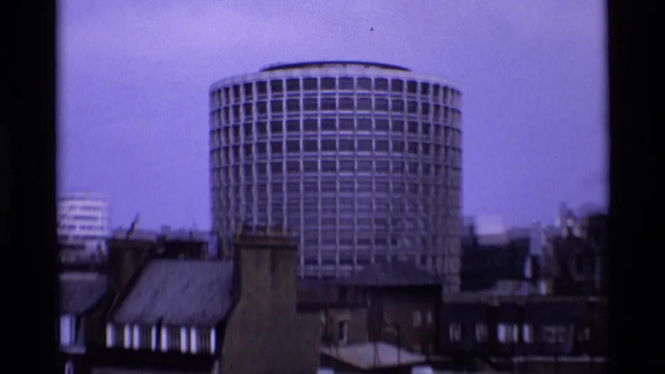 Circular modern London building seen above lower rooftops, filmed with a cool purplish 8mm cast.