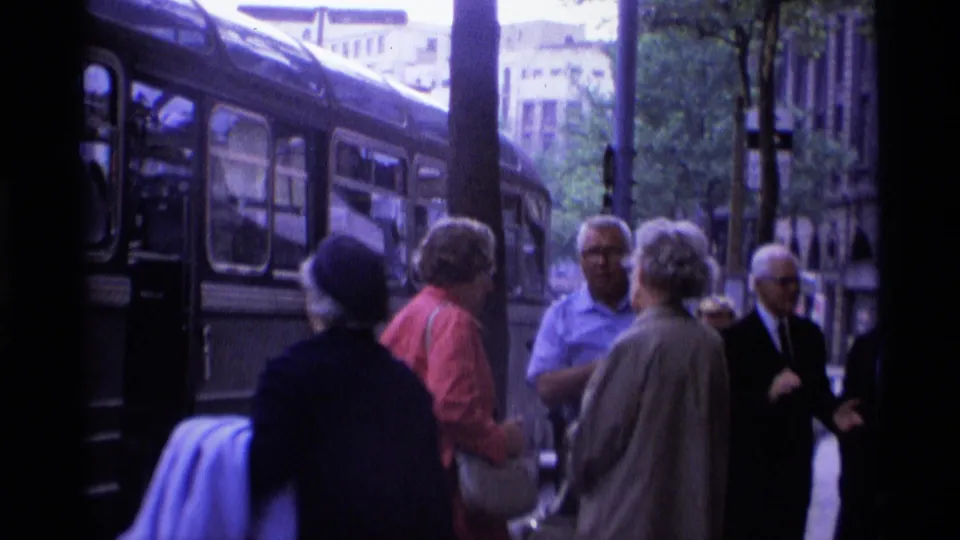Pedestrians in period clothing stand near a double-decker bus on a London sidewalk in 1969 home-movie footage.