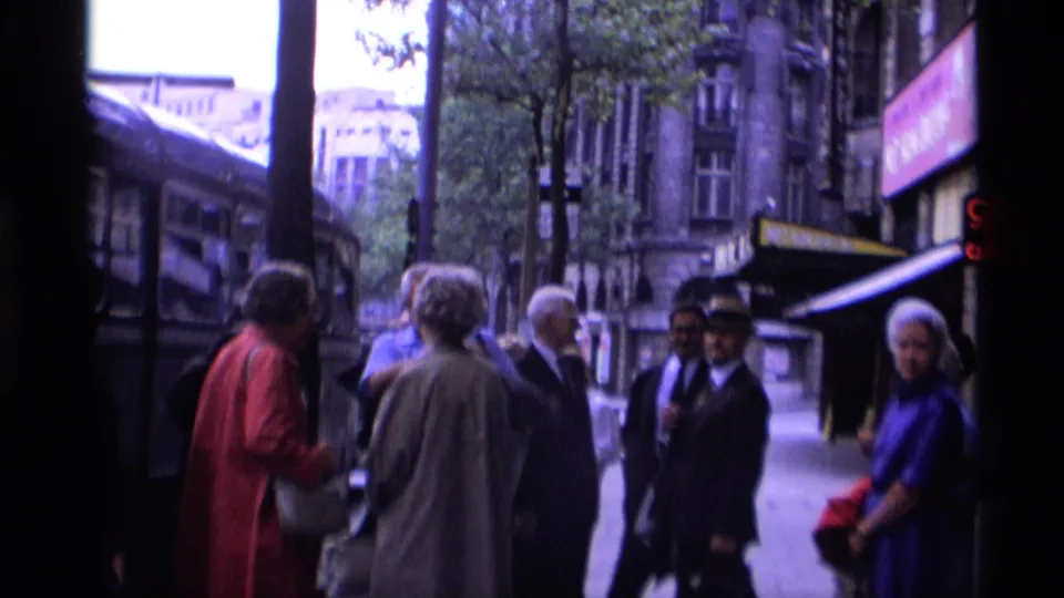 London pedestrians stand near a bus with shop awnings and older building facades visible along the street.