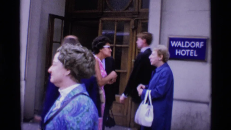 A group of adults gathers by the Waldorf Hotel entrance in London, including women in coats and a man in a suit.