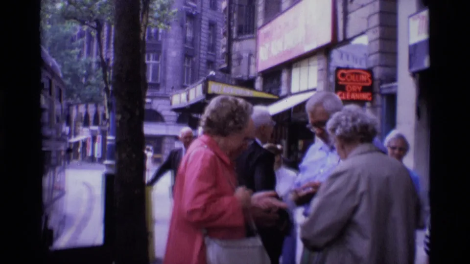 London street scene from 1969 showing adults gathered near shopfronts and a tree-lined pavement outside the hotel area.