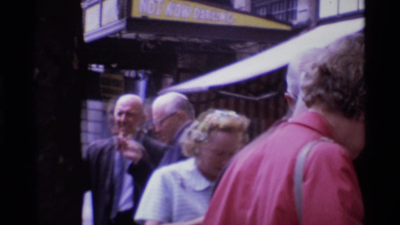 1969 London street frame with older adults talking and passing beneath a visible shop awning.