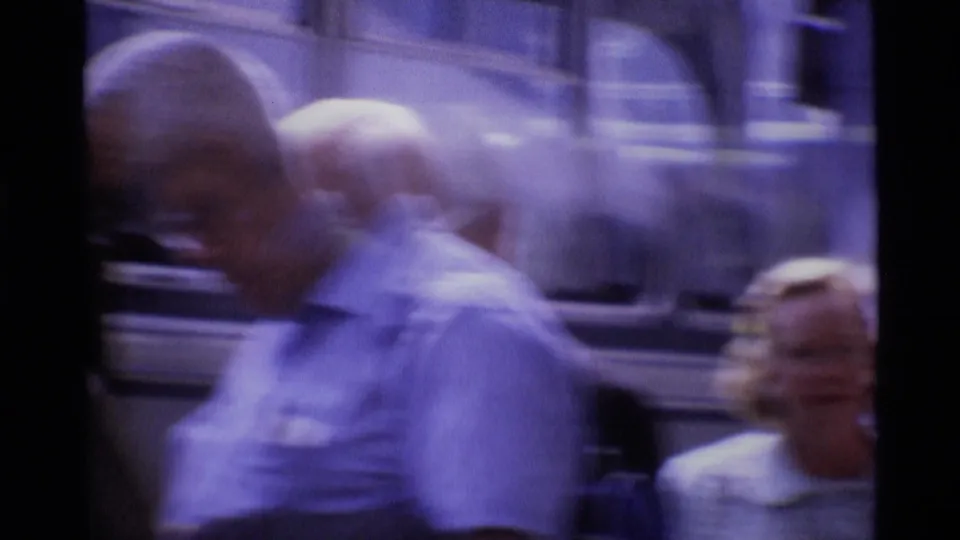1969 London 8mm frame with blurred pedestrians in front of a large vehicle and street-corner crowd.