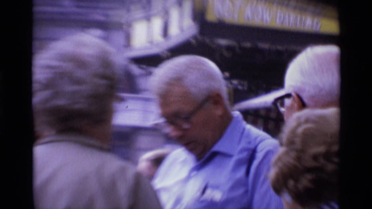 1969 London home-movie frame of older adults clustered on a downtown street, with glasses, white hair, and storefront signage visible.