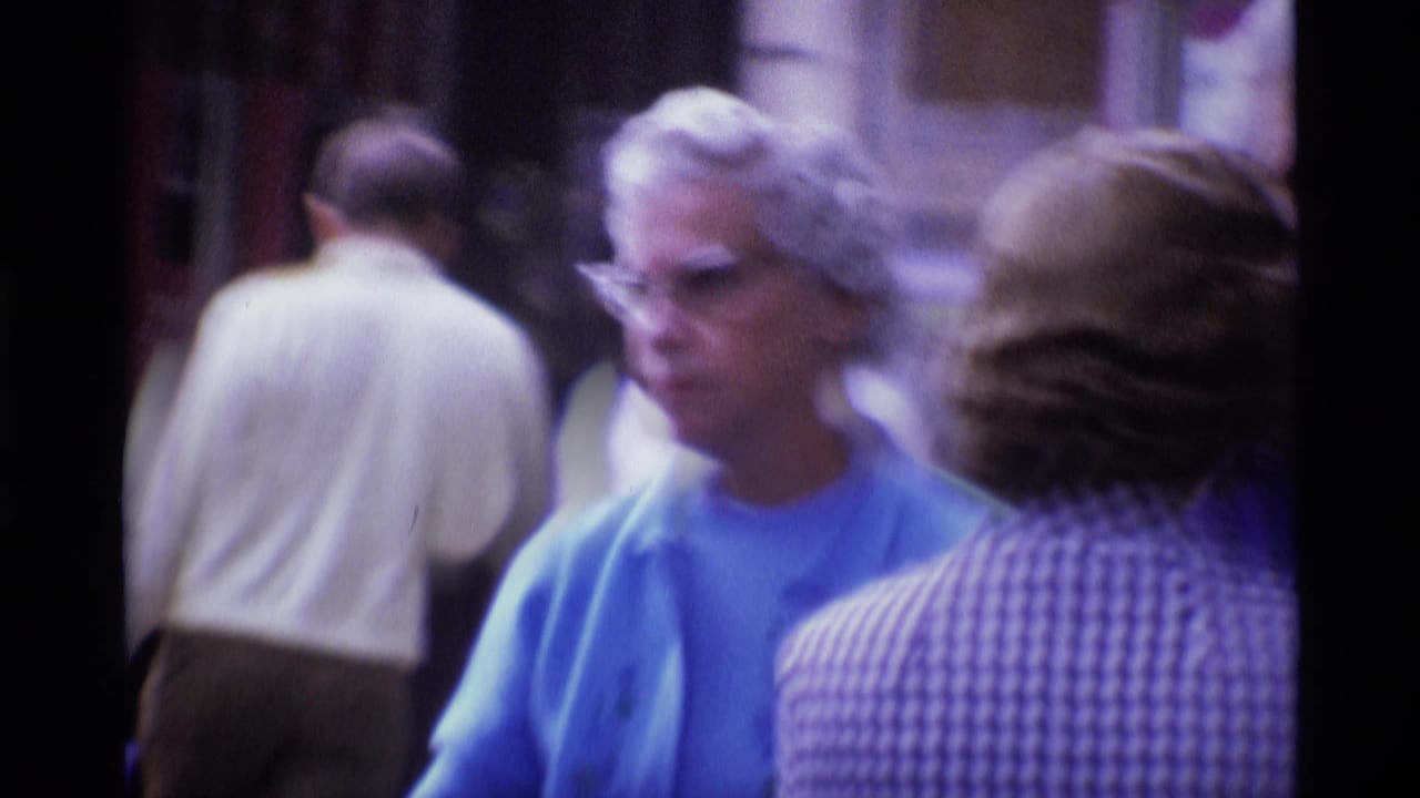1969 London street frame with a woman in a blue cardigan and glasses among pedestrians, seen in soft 8mm focus.