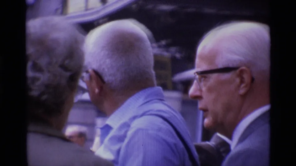 1969 London home-movie frame showing older men with white hair and glasses standing close together on a crowded street.