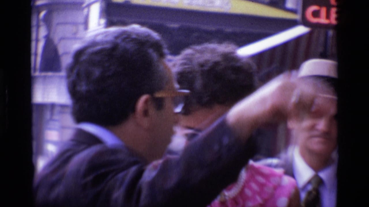 Suited man in glasses raises an arm near a woman in a pink polka-dot dress and a man in a hat.