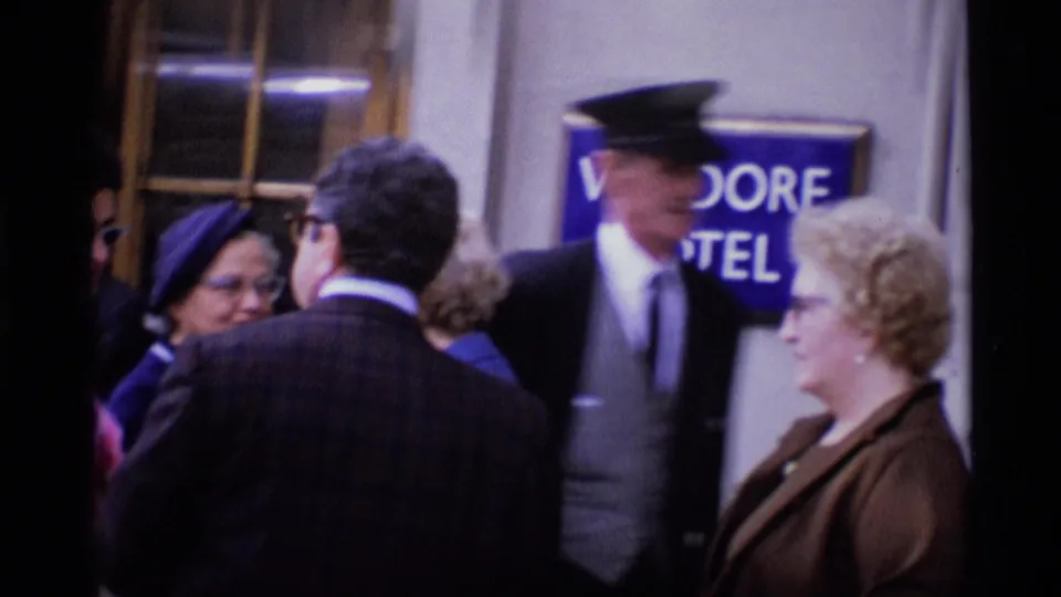 A crowded London hotel entrance shows a uniformed doorman, suited pedestrians, women in hats, and a partial Waldorf Hotel sign.