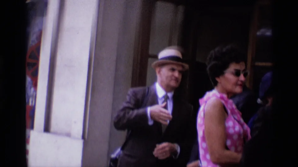 Near the London hotel entrance, a hat-wearing man and a woman in a pink polka-dot dress pass through a crowded doorway area.