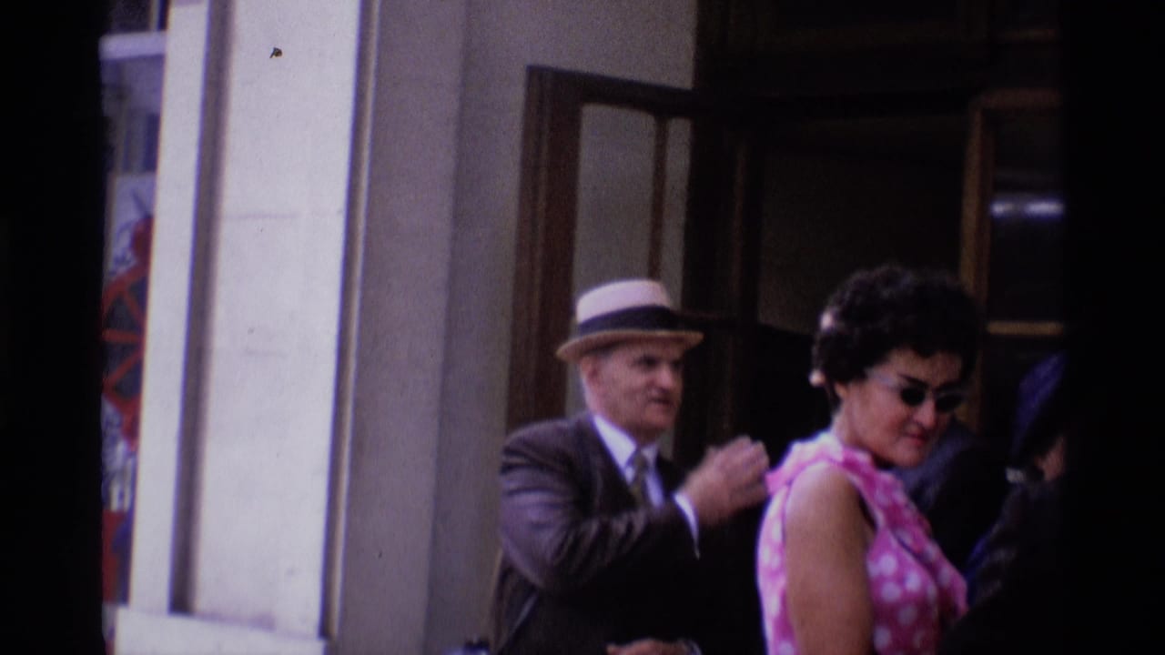 1969 London sidewalk scene with adults by a storefront, including a suited man in a hat and a woman in a pink patterned dress.