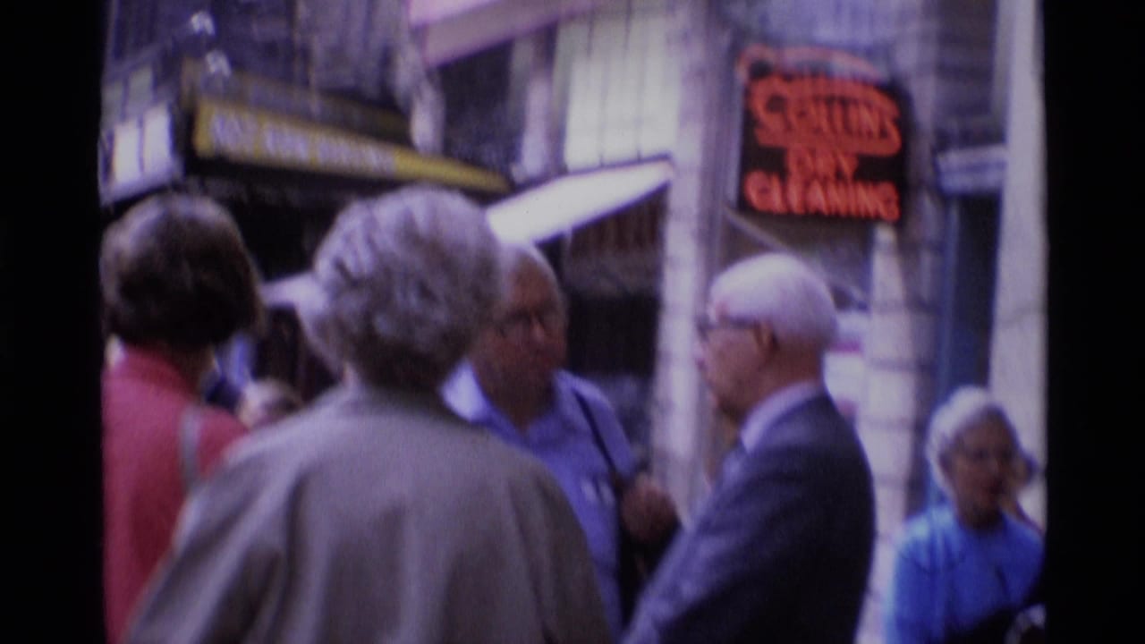 Adults converse on a sidewalk near storefronts and a red neon dry-cleaning sign.