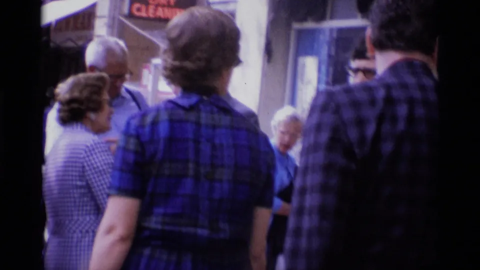 1969 London sidewalk crowd with adults in dresses and shirts moving past storefronts and street signage.