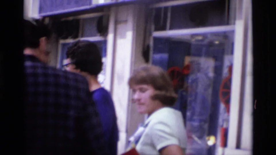 1969 London storefront view with a woman in a light coat, nearby pedestrians, reflective glass, and partial street signage.