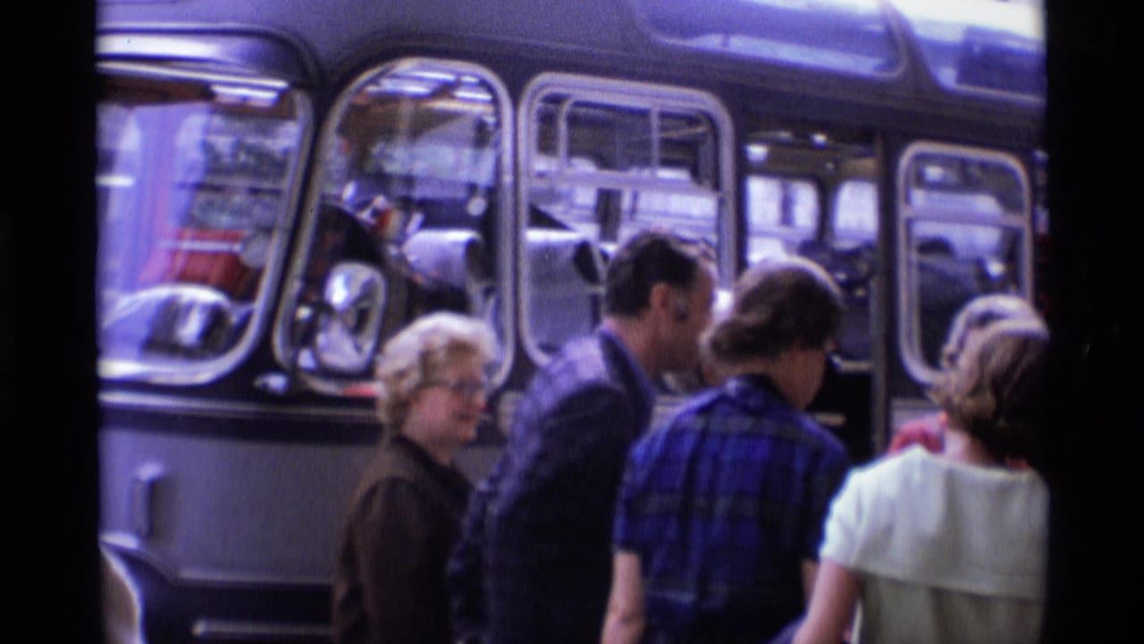 1969 London home-movie frame showing older travelers gathering beside a rounded-window coach before boarding.