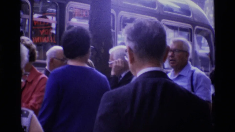 1969 London home-movie frame with older adults in sweaters and suits waiting beside a rounded-window coach.