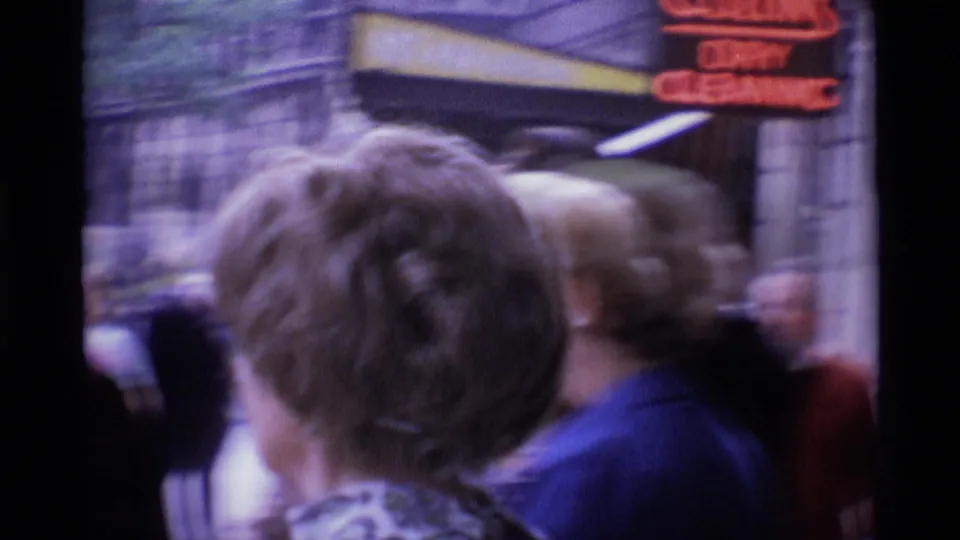 1969 London home-movie frame with blurred passengers, street signage, and the edge of a bus or storefront canopy.