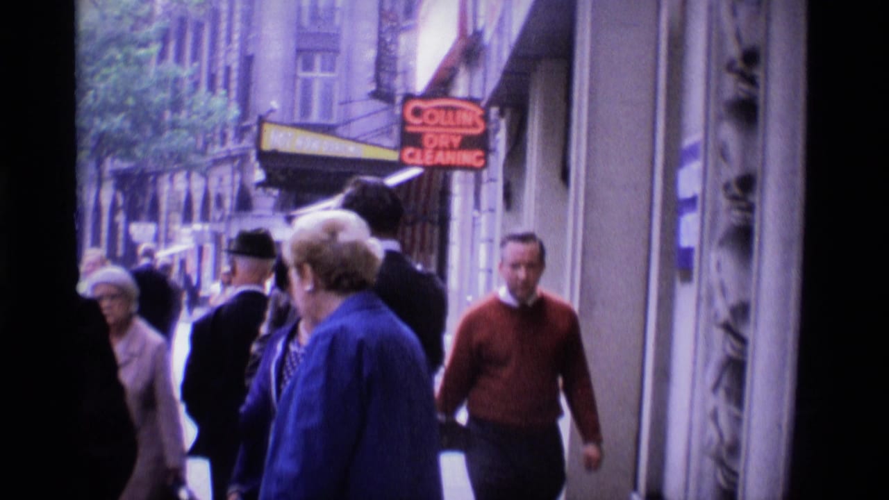 Pedestrians walk along a London sidewalk with a shop sign and city buildings visible.