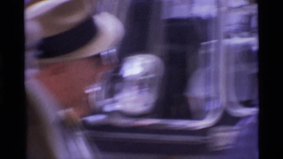 Blurred 1969 London home-movie frame with a brimmed hat and reflective metal fixtures in a crowded public setting.