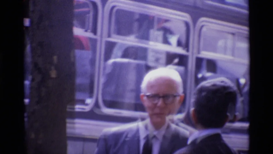 1969 London frame showing an older man in glasses near another adult, with large vehicle windows in the background.