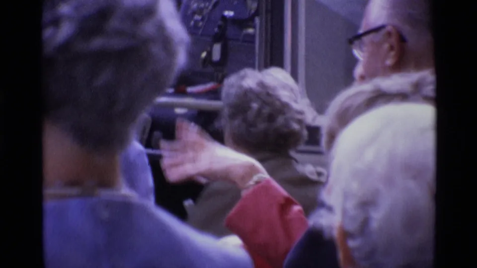 1969 London home-movie frame of adults in a crowded public area near vehicle-like fixtures and windows.