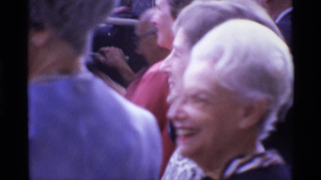 In 1969 London home-movie footage, an older woman smiles in soft focus while adults crowd closely around her.