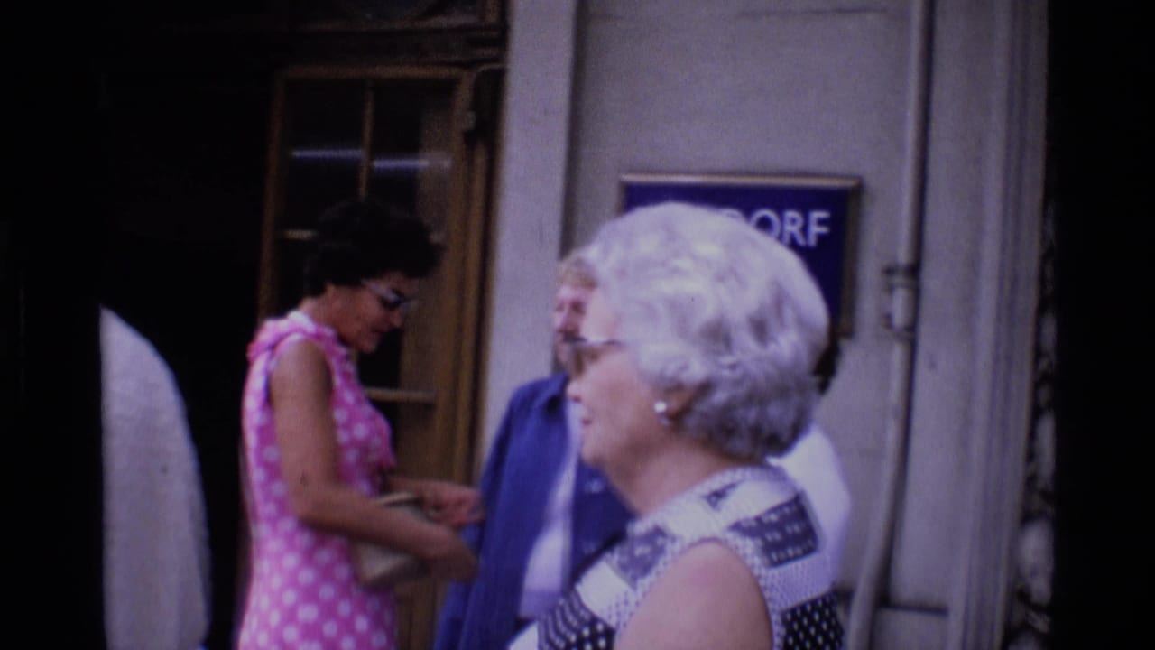Adults appear to greet one another near a doorway in London, with a woman in a pink polka-dot dress visible at center.