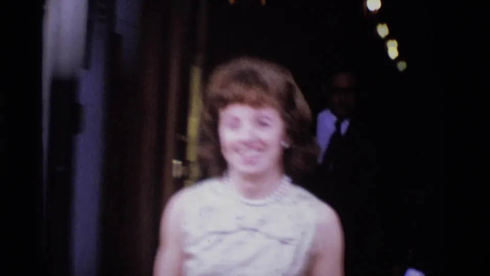A young woman in a light sleeveless dress smiles toward the camera near a dark entrance in the 1969 London scene.