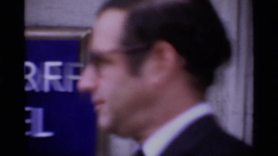 A suited man passes in soft-focus profile beside a blue sign with partial white lettering in the London gathering footage.