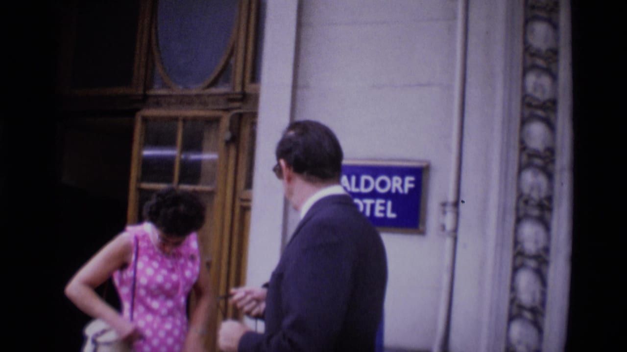 1969 London home-movie frame showing a woman in a pink patterned dress and a suited man near a hotel doorway and sign.