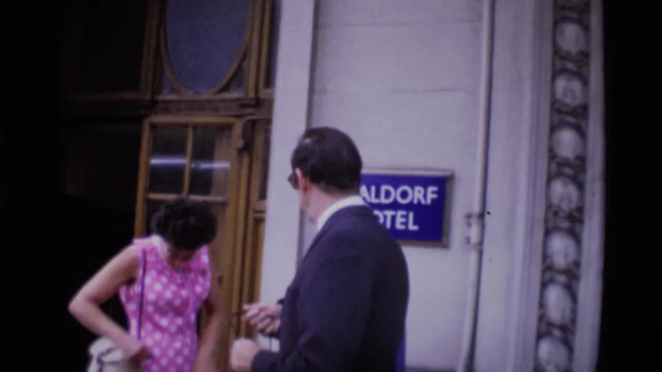 1969 London home-movie frame showing a woman in a pink patterned dress and a suited man near a hotel doorway and sign.
