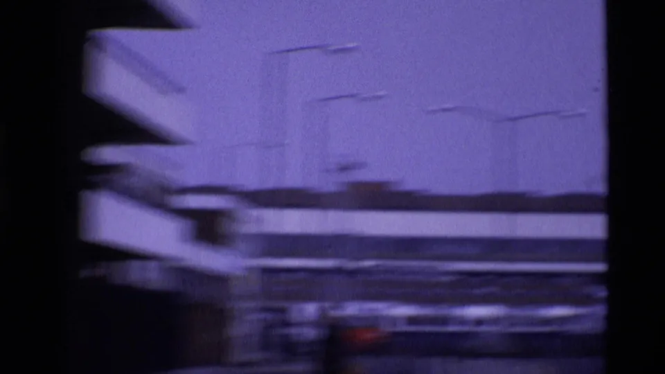 1969 London home-movie frame with a soft, blurred view of modernist architecture and street traffic.