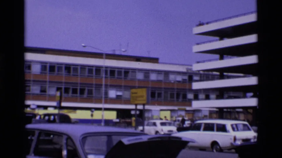 1969 London street scene with parked cars, pedestrians, a traffic light, and layered modern building facades.