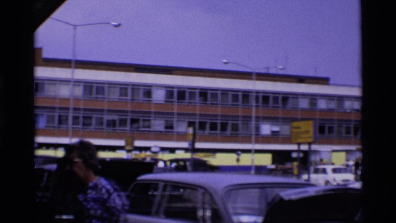 1969 London street scene with parked cars, a pedestrian in the foreground, and a large brick-and-glass building partly obscured by the filming position.