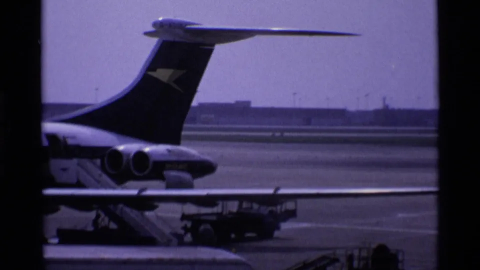 1969 London airport home-movie frame showing a parked aircraft tail beyond terminal window mullions and tarmac equipment.