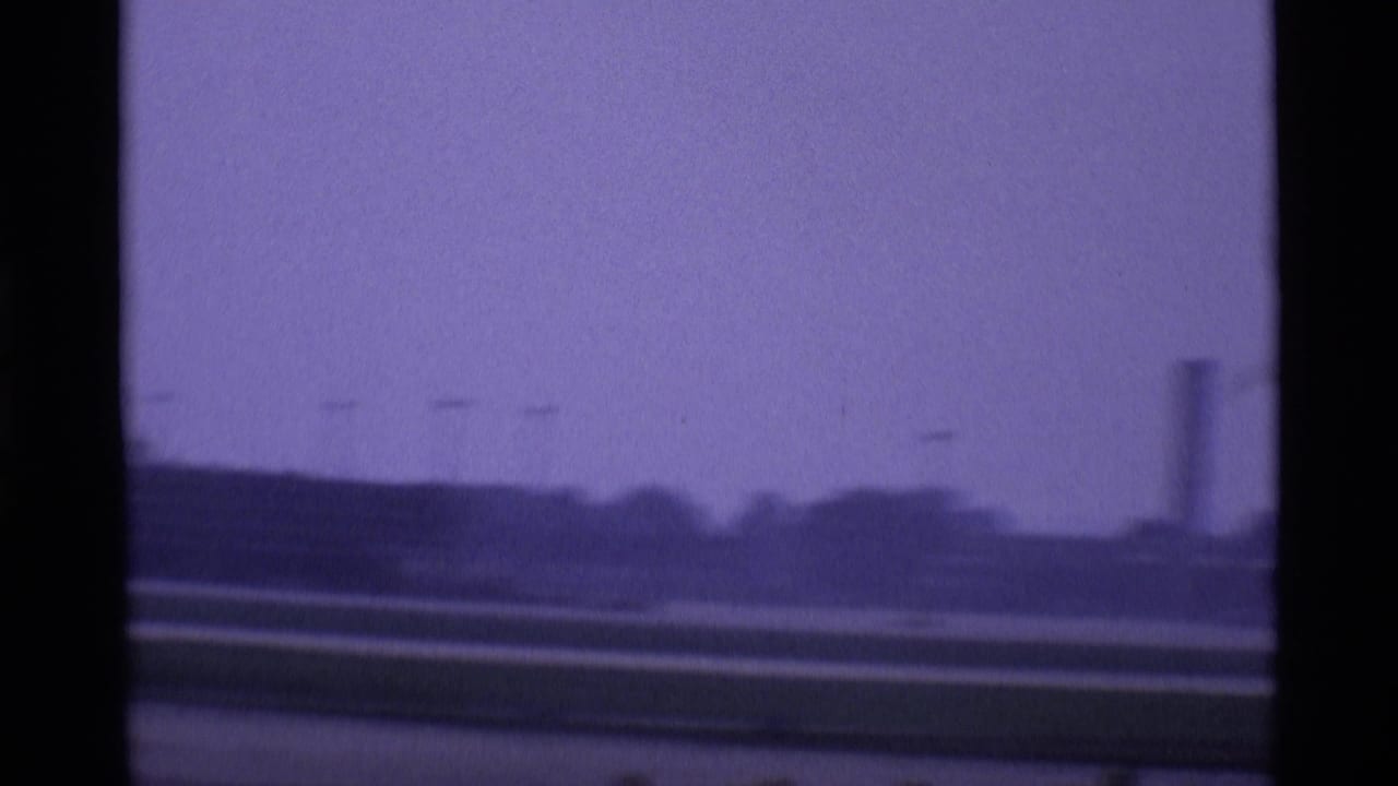 1969 London airport view across runway and distant airport structures, divided by dark terminal window framing.
