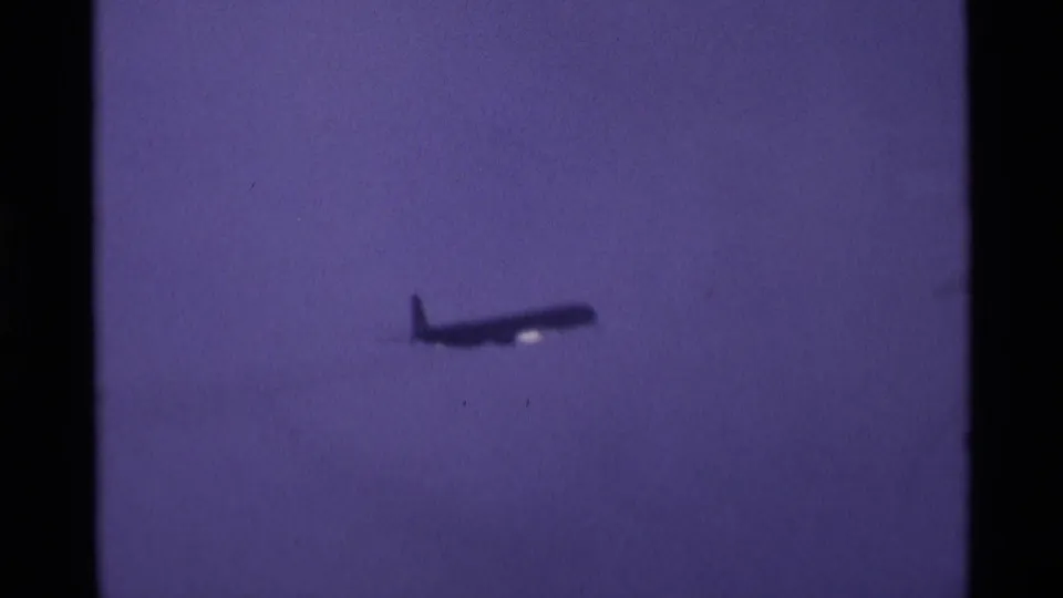 A small distant airliner silhouette crosses the hazy sky in this 1969 London airport 8mm frame.