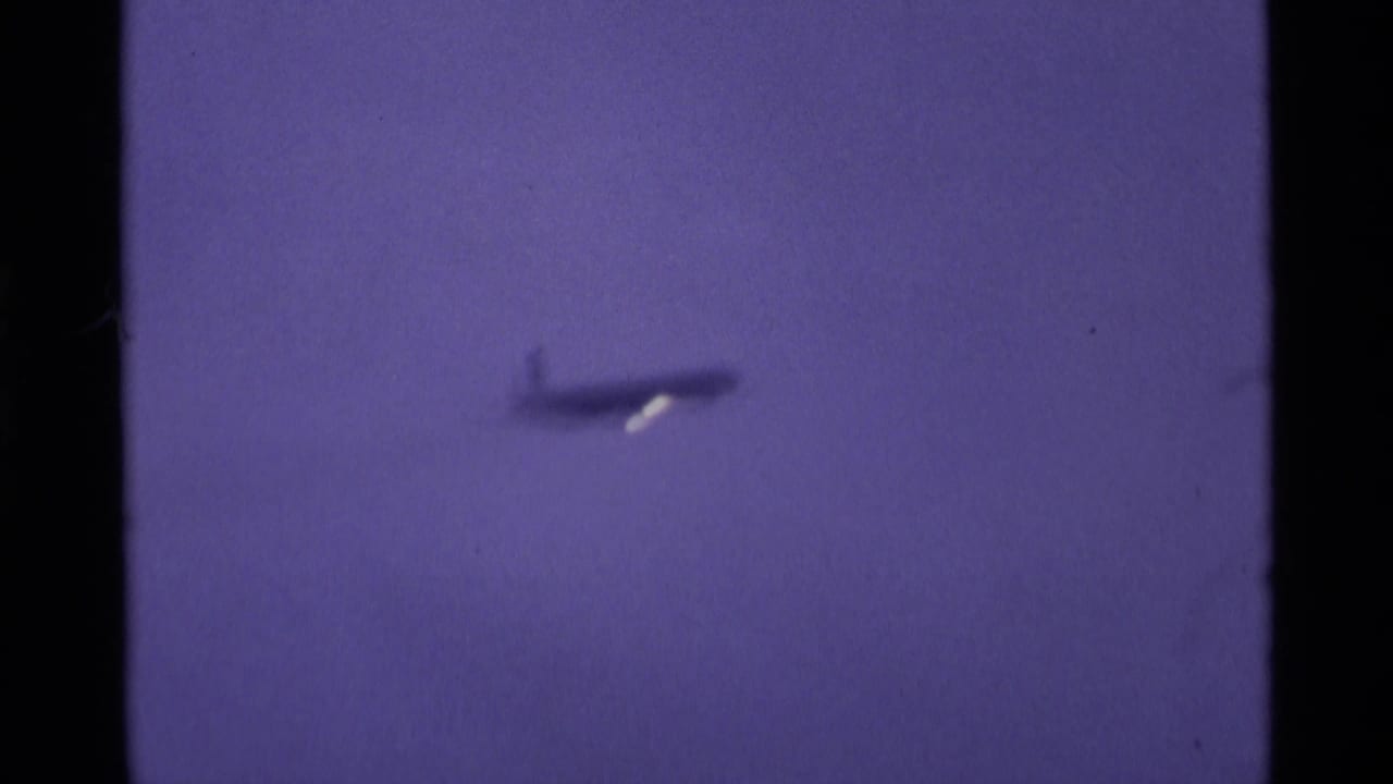 A small airliner silhouette appears high in a purple-hued London sky in this 1969 8mm home-movie frame, with faint haze behind it.