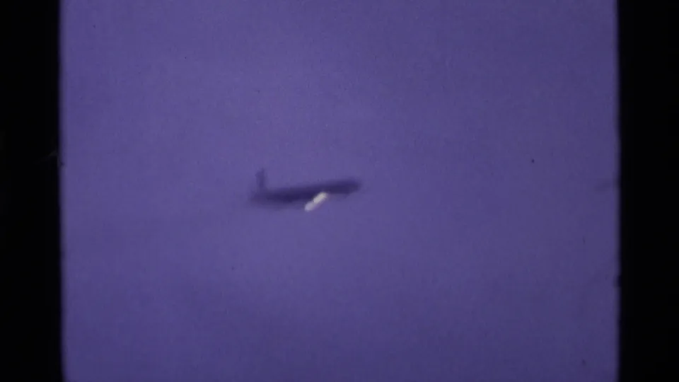 A small airliner silhouette appears high in a purple-hued London sky in this 1969 8mm home-movie frame, with faint haze behind it.