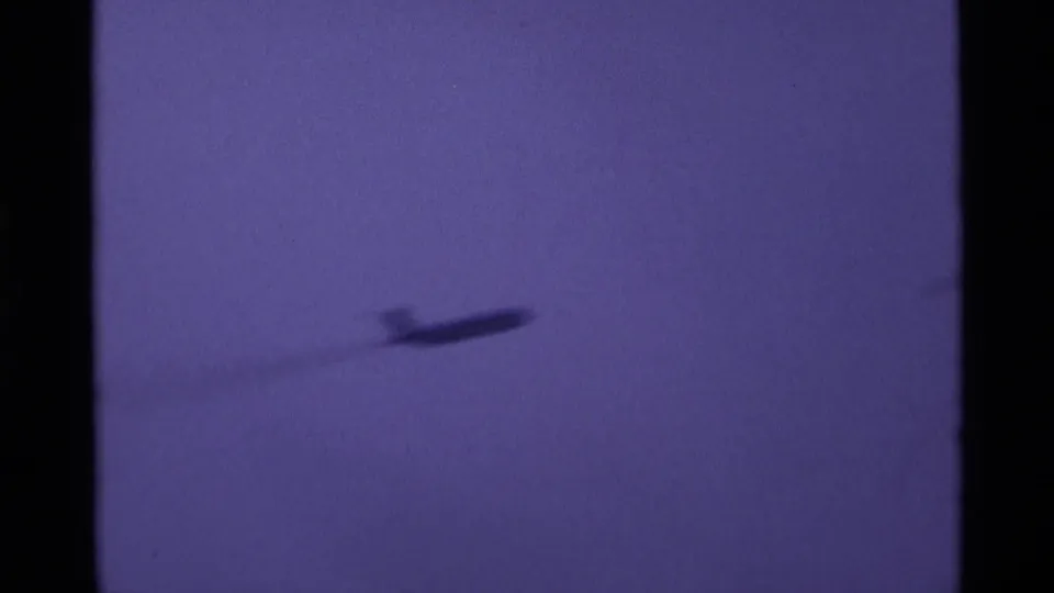 The aircraft remains a dark, distant shape against the purple cast of the 1969 London home-movie sky, with slight blur from motion and film texture.