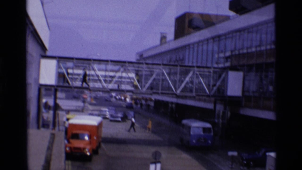 1969 London airport terminal approach with an enclosed pedestrian bridge, period vehicles, and small travelers or staff moving near the roadway.