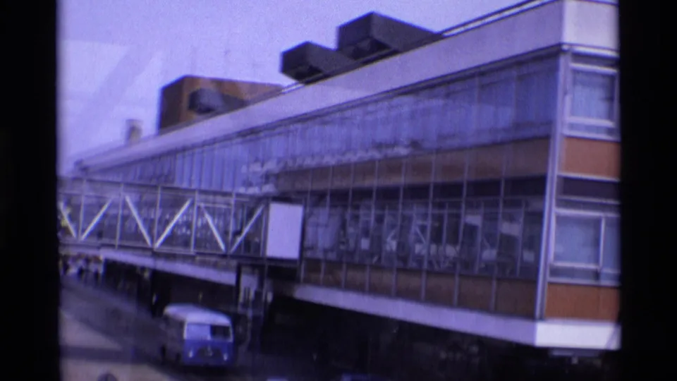 1969 London airport exterior showing the terminal wall, glass-fronted pedestrian bridge, blank sign panels, and a blue service or passenger van.
