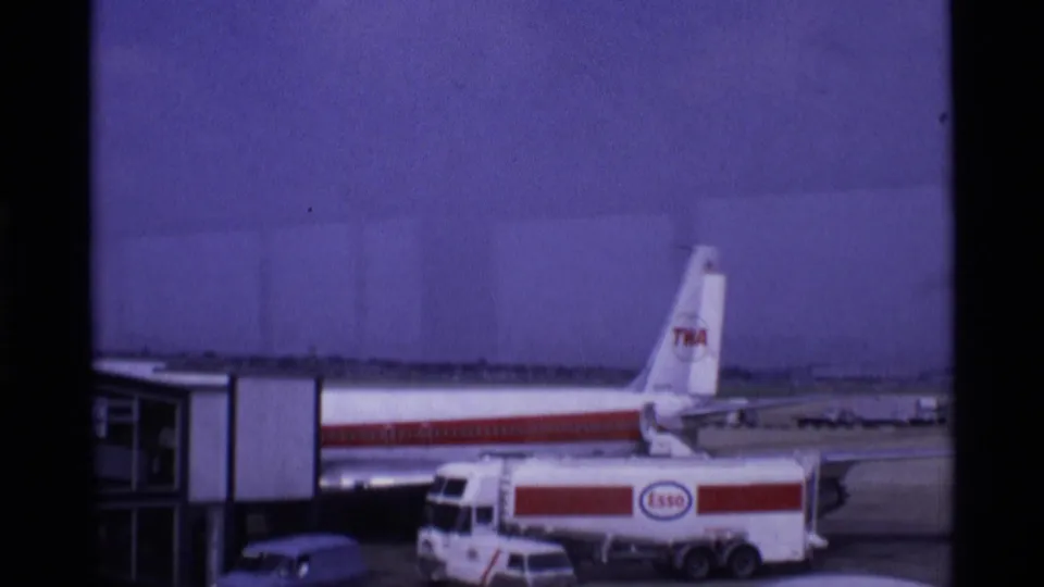 1969 London airport apron with a TWA-marked aircraft at the terminal and an Esso fuel tanker positioned near service vehicles.