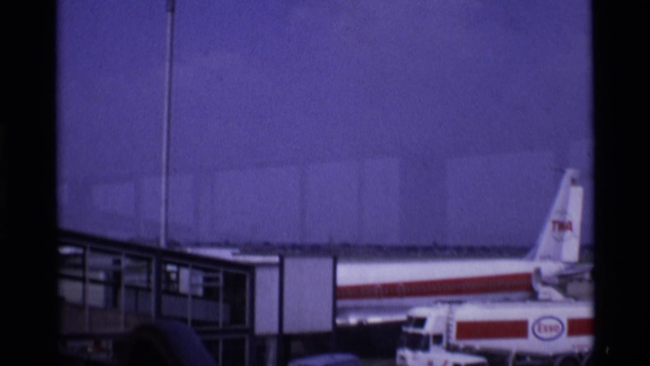 1969 London airport footage showing a TWA jet tail and fuselage beside a gate structure, with ground vehicles in the foreground.
