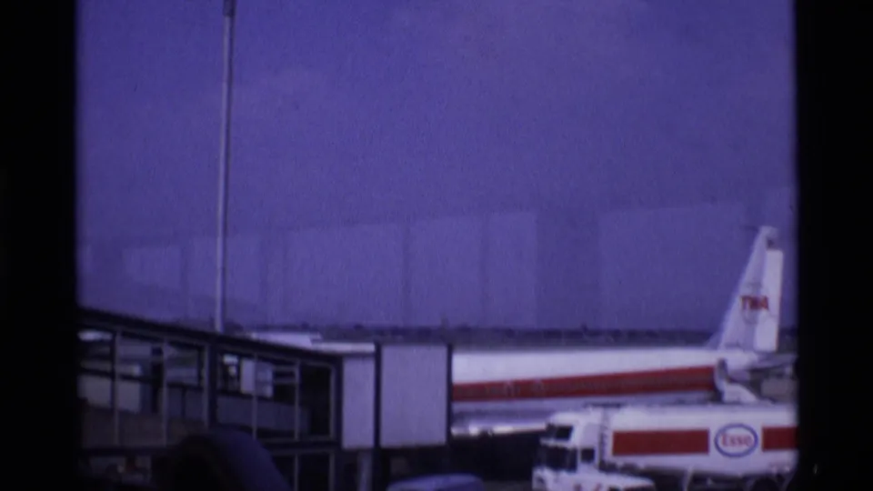 1969 London airport footage showing a TWA jet tail and fuselage beside a gate structure, with ground vehicles in the foreground.