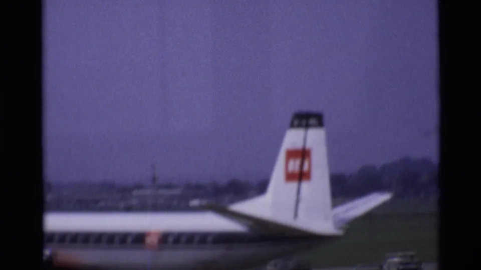 1969 London airport view of a BEA aircraft tail and rear fuselage on the ramp, with distant airfield structures beyond.