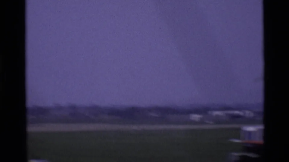 Soft, grainy 1969 London airport tarmac view with distant low buildings and a partial aircraft edge in frame.